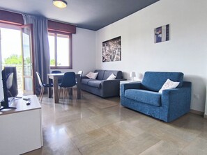 A modern living room with a blue armchair, grey sofa, and a dining area with a table and chairs. - Cozy studio with balcony (Lido di Jesolo)