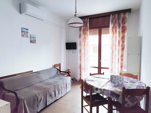 A room with a sofa, a dining table with chairs, a ceiling fan, and a window with curtains. - Seaside apartment with balcony (Lido di Jesolo)
