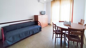 Una habitación pequeña y bien iluminada con un sofá, una mesa con sillas, un televisor sobre un mueble y una ventana con cortinas.
