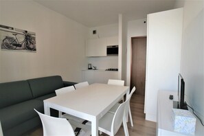 A modern living space with a sofa, a dining table, and chairs. There is a kitchen area with cabinets and a microwave. A television is mounted on the wall. - Comfortable apartment in Jesolo (Lido di Jesolo)