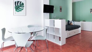 Quarto moderno com mesa e cadeiras de vidro, escrivaninha branca com TV de tela plana, cama com parede verde e ar condicionado instalado no teto.