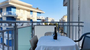 Een balkon met een tafel gedekt voor twee personen, een fles wijn en twee glazen. Het balkon kijkt uit op een rij gebouwen met balkons.