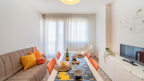Een moderne woonkamer met een eettafel voor vier personen, een bank met oranje kussens, een flatscreen televisie en een groot raam met dunne gordijnen.