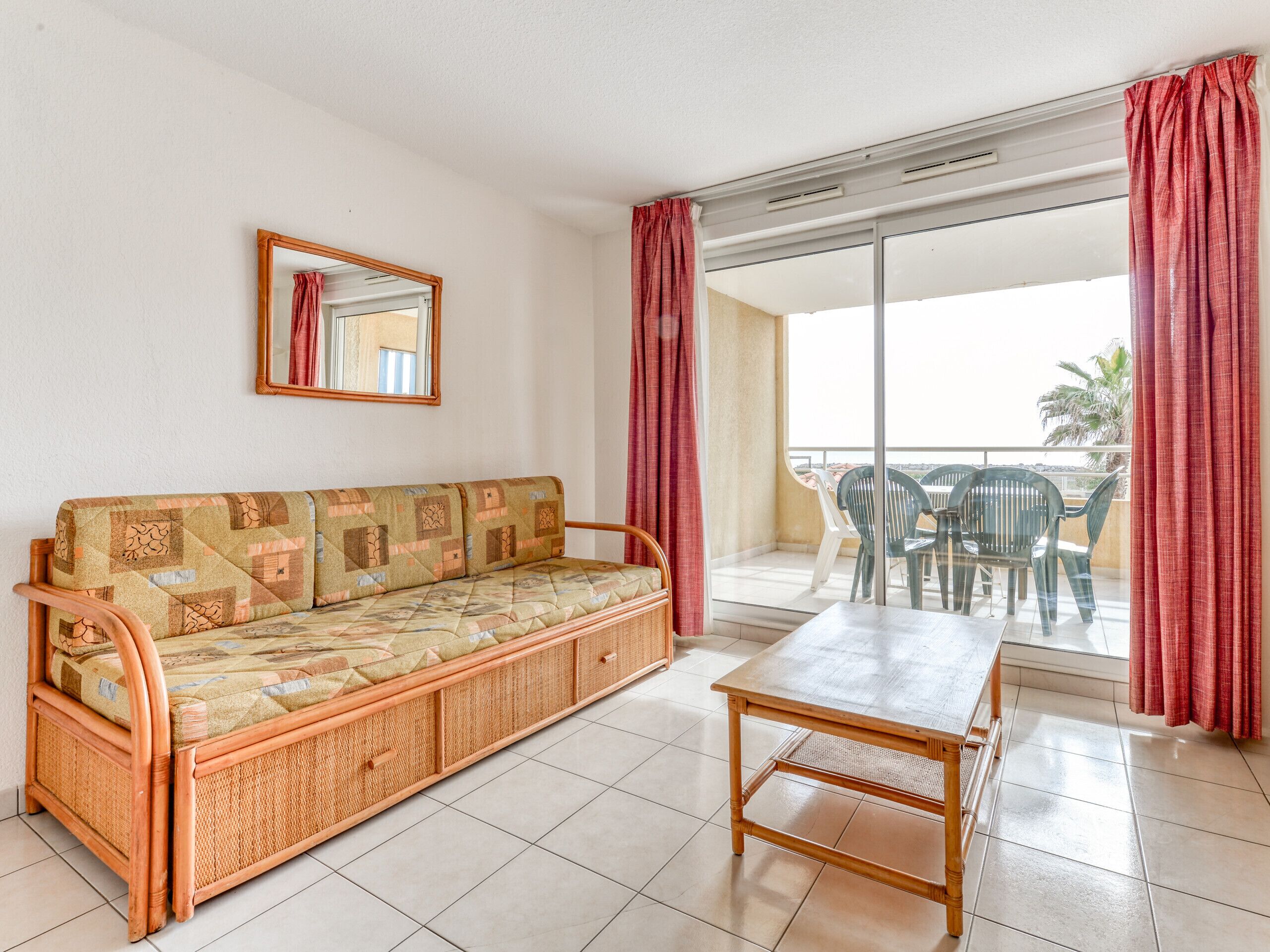 A room with a sofa, a table, a mirror, and a balcony with chairs.