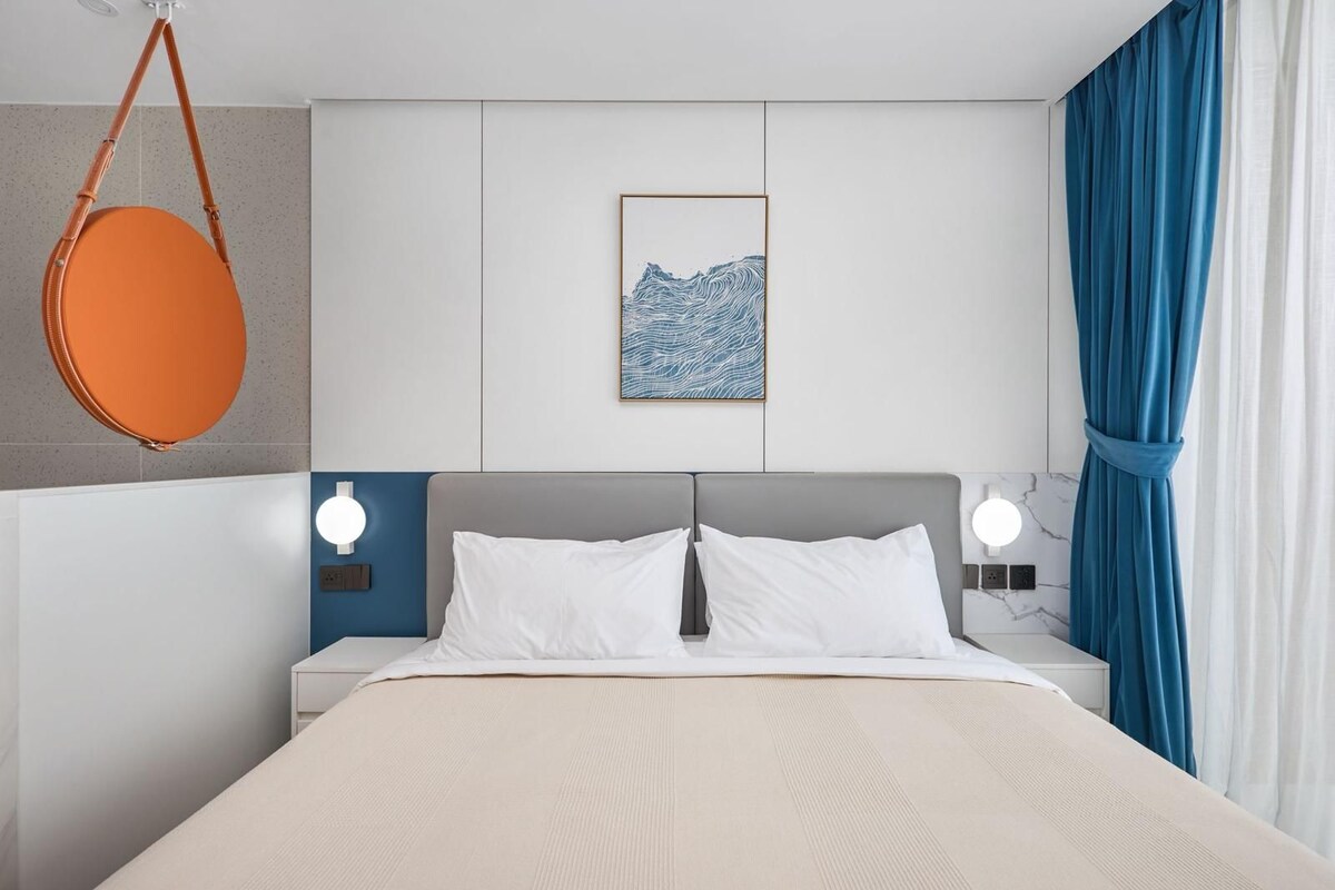 Ein Hotelzimmer mit einem Bett, zwei Nachttischen, einer hängenden orangenen Tasche, einem gerahmten Kunstwerk und einem Fenster mit Vorhängen.