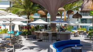An outdoor bar area with seating and umbrellas, surrounded by palm trees and modern architecture.