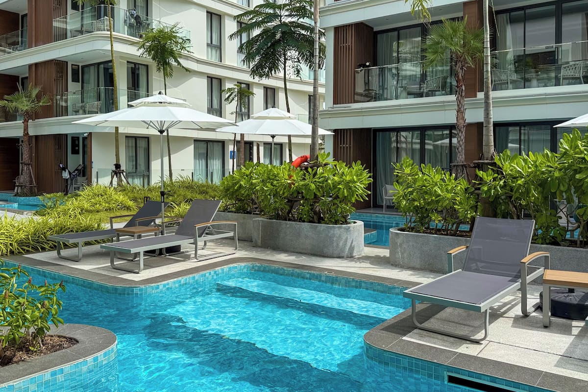 A swimming pool with lounge chairs and umbrellas, surrounded by greenery and a modern building with balconies.