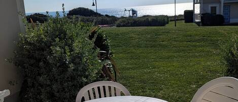 A patio with a round table and white plastic chairs, overlooking a grassy area and a body of water.