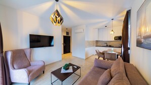 A modern living room with a sofa, armchair, and coffee table. There is a flat-screen TV mounted on the wall, a kitchen area with cabinets and a microwave, and a dining table with chairs. - Spacious 1BR Apt for 3 w/ Terrace & Pool (Sutomore)