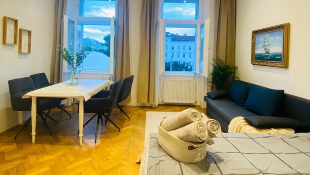Une chambre d’hôtel moderne avec un parquet en bois, une table à manger avec des chaises, un canapé et une grande fenêtre donnant sur des immeubles et des arbres.