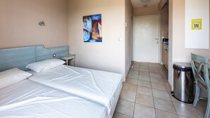 Quarto de hotel com cama, uma mesa pequena, um cesto de lixo, um quadro na parede e uma porta que dá para uma cozinha compacta.