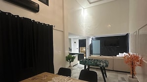 Un coin repas avec une table en bois, des chaises noires et une nappe en dentelle. On aperçoit au fond un baby-foot, une plante en pot et un luminaire suspendu.