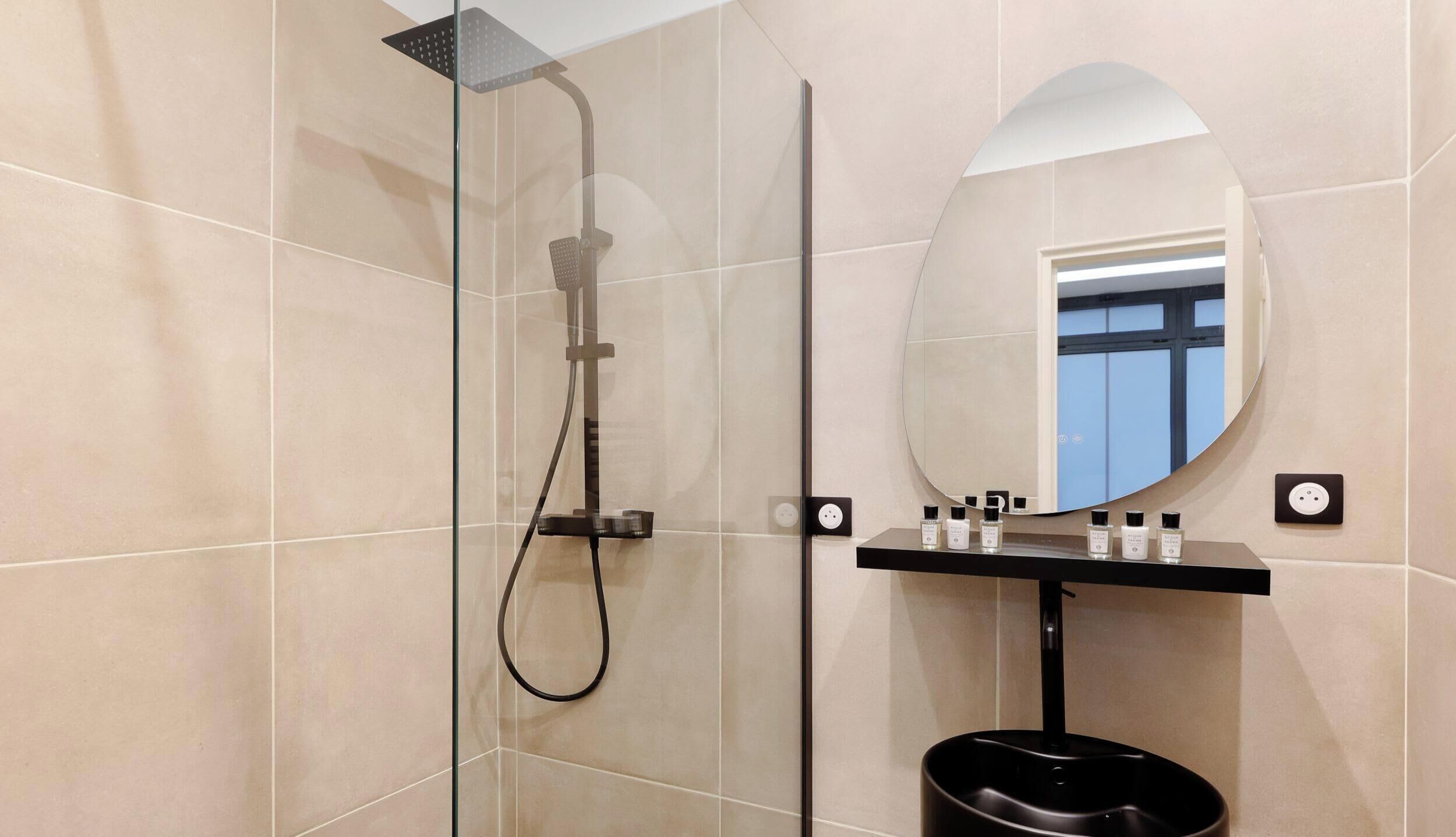 Un baño moderno con ducha envuelta en vidrio, espejo redondo y lavamanos negro con artículos de aseo.