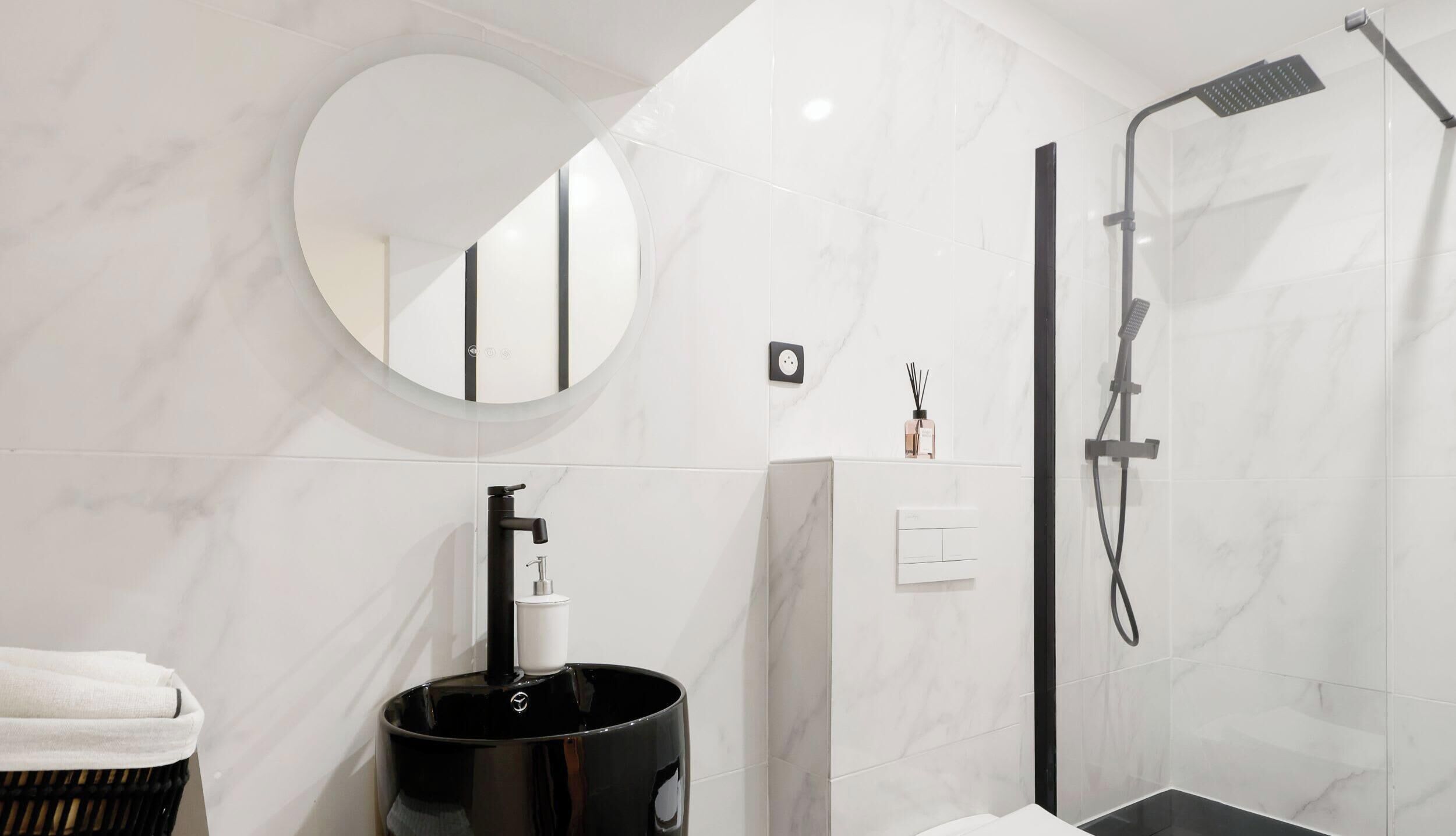 Un baño moderno con un espejo redondo, un lavabo negro, cabina de ducha de vidrio y pared revestida de azulejos blancos.