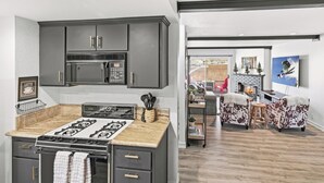 A modern kitchen with a gas stove, dark cabinets, and a microwave. There is a dining area with a fireplace and a large window. - Ski In Ski Out Condo at Bear Mountain! HOT TUB! Walk Onto Chair Lift 9! (Big Bear Lake)