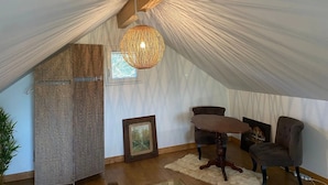 Ein Zimmer mit schräger Decke, einer gewobenen Leuchte, einem gerahmten Bild, einem dunklen Ohrensessel und einem kleinen Tisch.