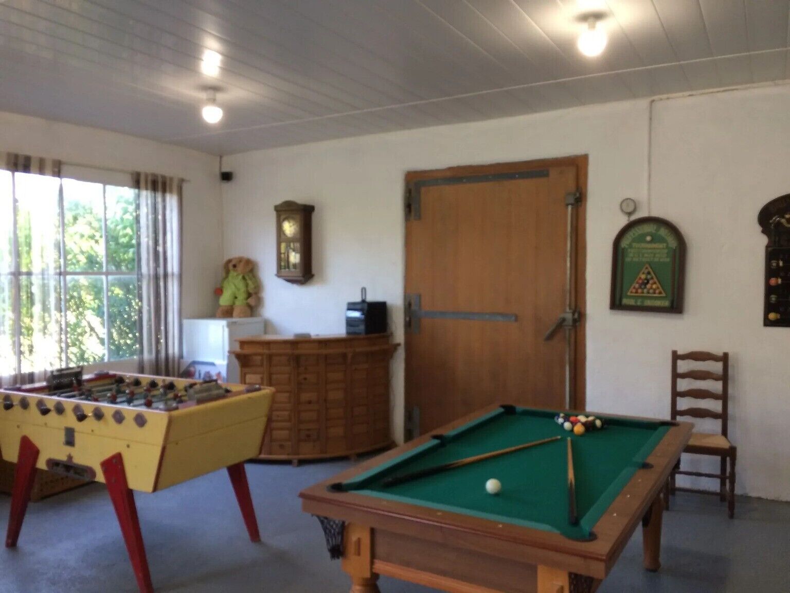 Una stanza con tavolo da biliardo, tavolo da calcio, un mobile in legno, una sveglia e una finestra con tende.