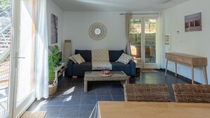 Een woonkamer met een bank, salontafel en een eettafel. Er is een groot raam met gordijnen, een wandspiegel en een houten bijzettafeltje.