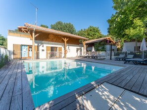 Una piscina con terraza de madera, zona de estar cubierta y un edificio con tejado de baldosas.