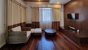 A room with a wooden ceiling, a flat-screen TV mounted on the wall, a sofa, a small ottoman, and a window with curtains. - Amatapura Beach Villa 14 (Krabi)