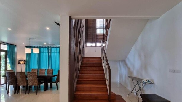 Un interior moderno con una escalera de madera que conduce a un nivel superior, una zona de comedor con sillas y una mesa, y una mesita auxiliar con una tabla de planchar.