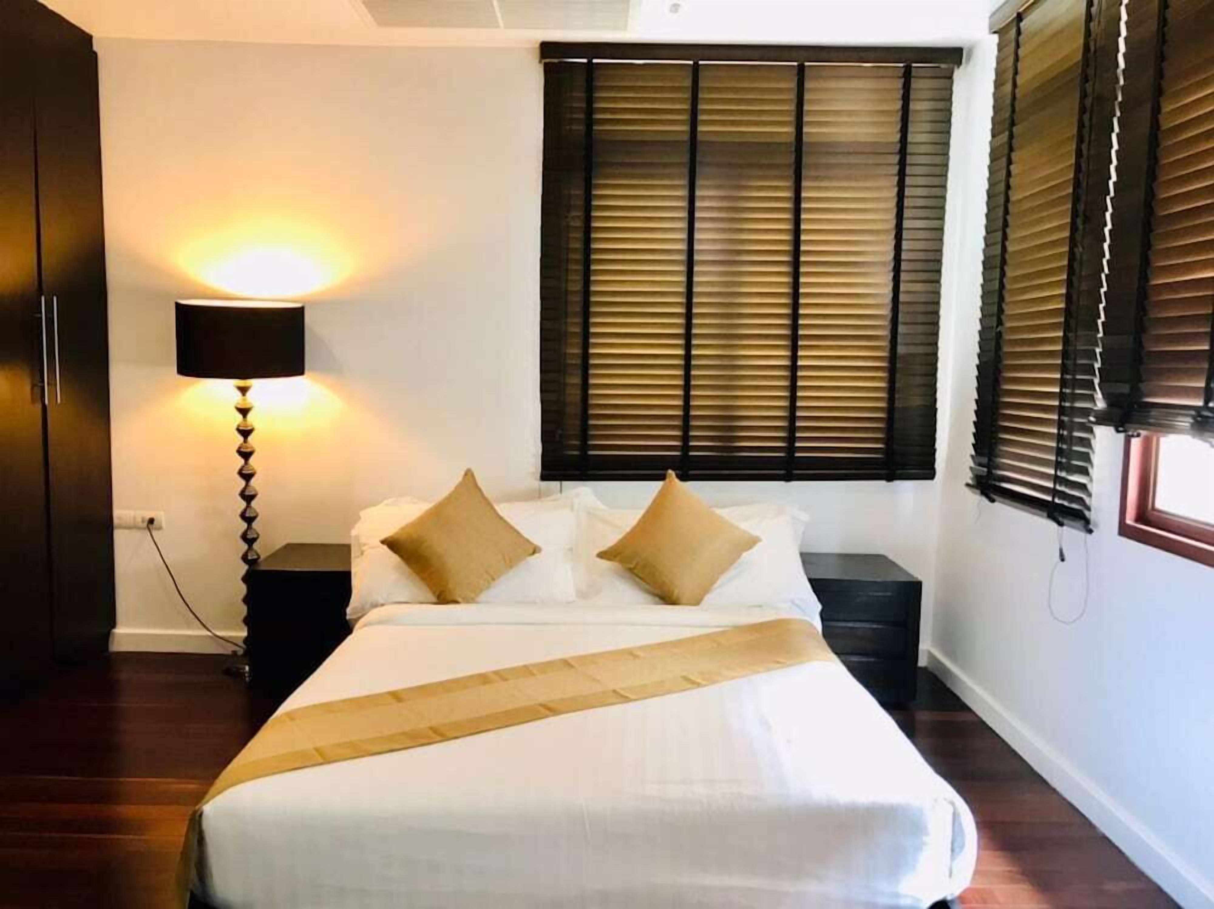 A hotel room with a bed, a bedside lamp, a window with blinds, and a wooden floor.