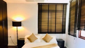Habitación de hotel con cama, lámpara de noche, ventana con persianas y suelo de madera.