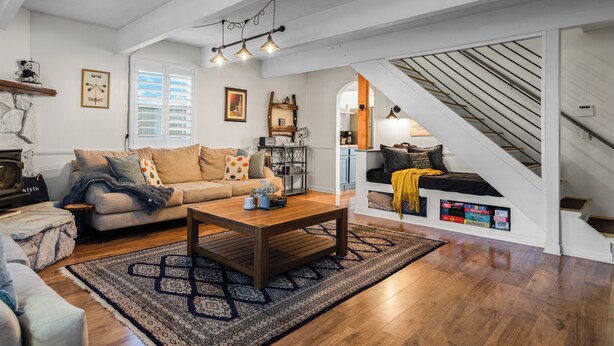 Un salón amplio con una mesa de centro de madera, un sofá color beige y una alfombra estampada. Una escalera con barandilla de madera conduce a un nivel superior.