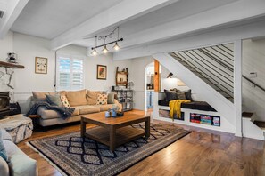 Ein geräumiges Wohnzimmer mit einem Holztisch, einem beigen Sofa und einem gemusterten Teppich. Eine Treppe mit Holzgeländer führt auf eine höhere Ebene.
