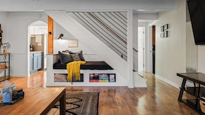 Una sala moderna con piso de madera, un sofá negro con una manta amarilla, una escalera y un televisor de pantalla plana montado en la pared.