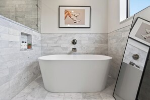 A modern bathroom with a freestanding bathtub, marble walls, a framed artwork, and a small shelf with toiletries. - Book for ACL | Lake Austin Views | Pool, Firepit + Game Room | Sundown Social (Austin)