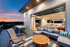 A modern outdoor deck with a dining area, seating, and a view of the interior. - Book for ACL | Lake Austin Views | Pool, Firepit + Game Room | Sundown Social (Austin)