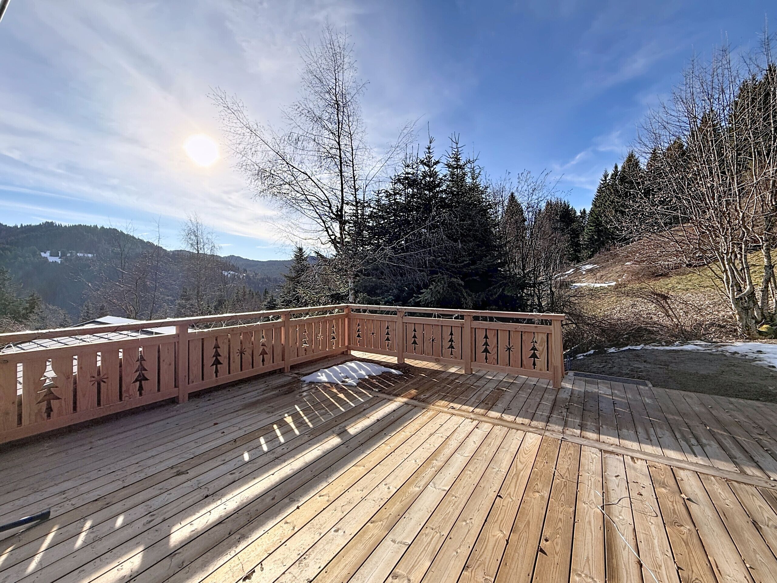A wooden deck with a railing, surrounded by snow-covered ground and trees, overlooking a mountainous landscape.