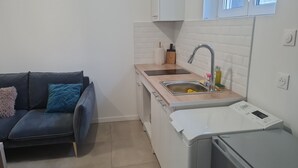 Una cocina compacta con un lavamanos, una encimera y un lavavajillas. En la zona de estar contigua hay un sofá oscuro con cojines.