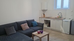 Un salon moderne avec un canapé gris foncé, une petite table basse en bois et un coin cuisine doté de meubles blancs, d’un évier et d’un lave-vaisselle compact.