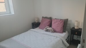 Una cama bien hecha con un edredón blanco, dos almohadas rosas y una almohada gris. Se observa una mesita de noche con una lámpara y un objeto decorativo.