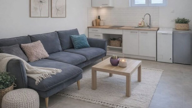 Una sala moderna con un sofá gris, una pequeña mesa de centro de madera, y una cocina con armarios blancos y una ventana sobre el fregadero.