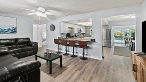 Una sala de estar moderna con un sofá de cuero negro, una mesa de centro y una zona de bar con taburetes. En el fondo, se ve una cocina con un refrigerador de acero inoxidable y una zona de comedor con vista a la piscina.