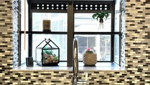 Una cocina con un amplio ventanal, un lavamanos de acero inoxidable y una estantería decorativa con plantas y un reloj.
