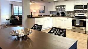Una cocina moderna con armarios blancos, un microondas, un horno y una cafetera. Hay una mesa de comedor con sillas y un cuenco decorativo.