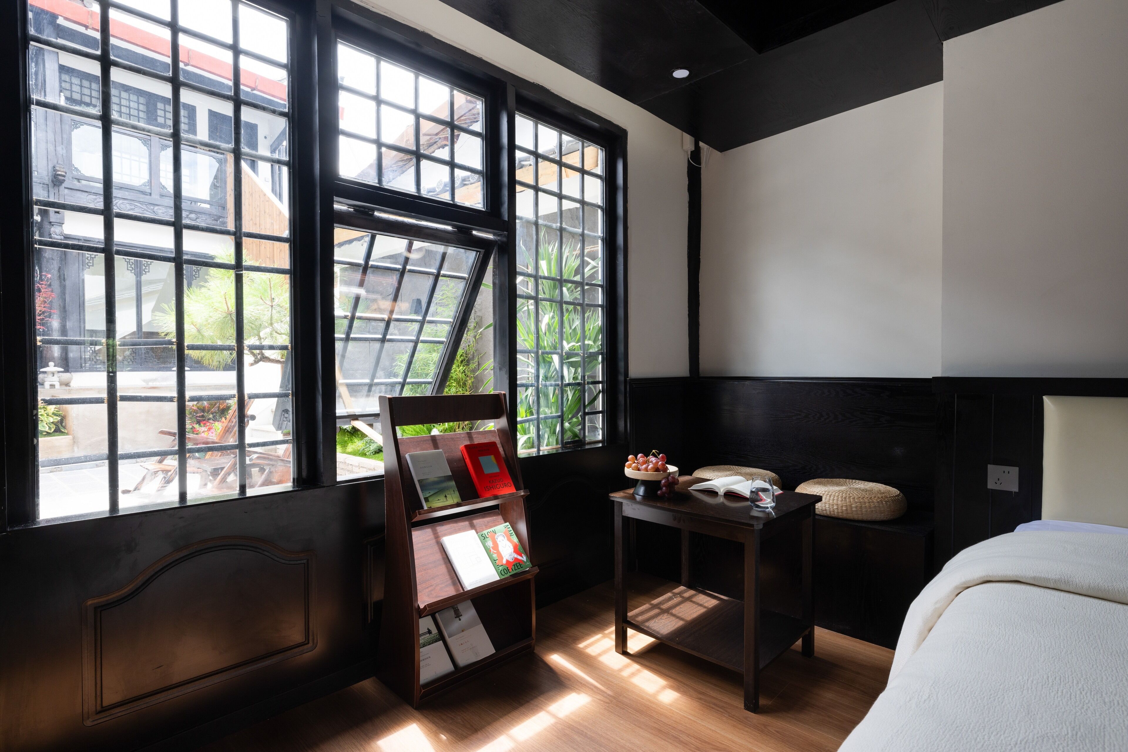 Ein Zimmer mit großem Fenster, Nachttisch mit Büchern, Obstschale und einem Bett.