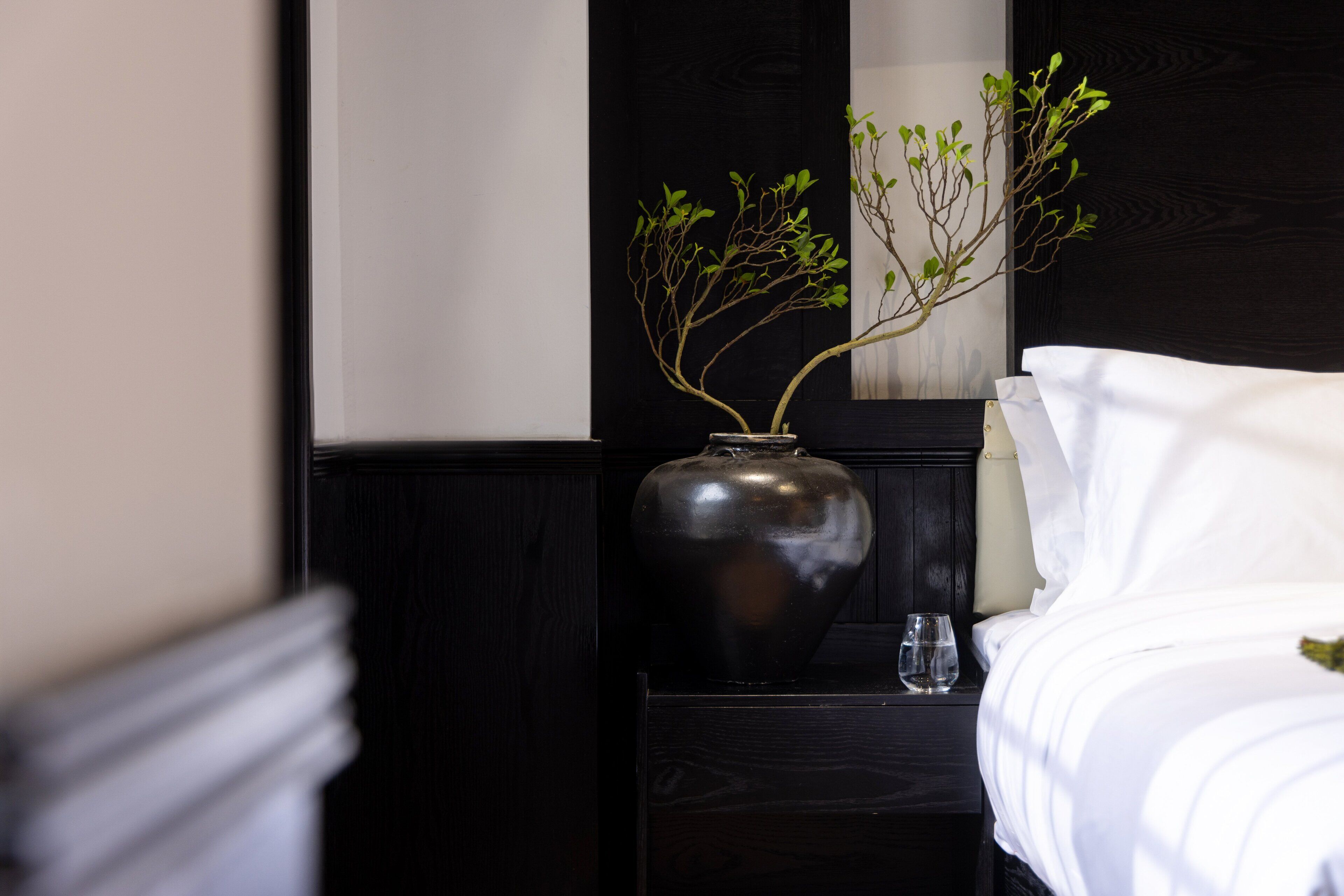 A neatly made bed with white linens, a dark wooden nightstand with a vase containing branches and a glass, and a black headboard.