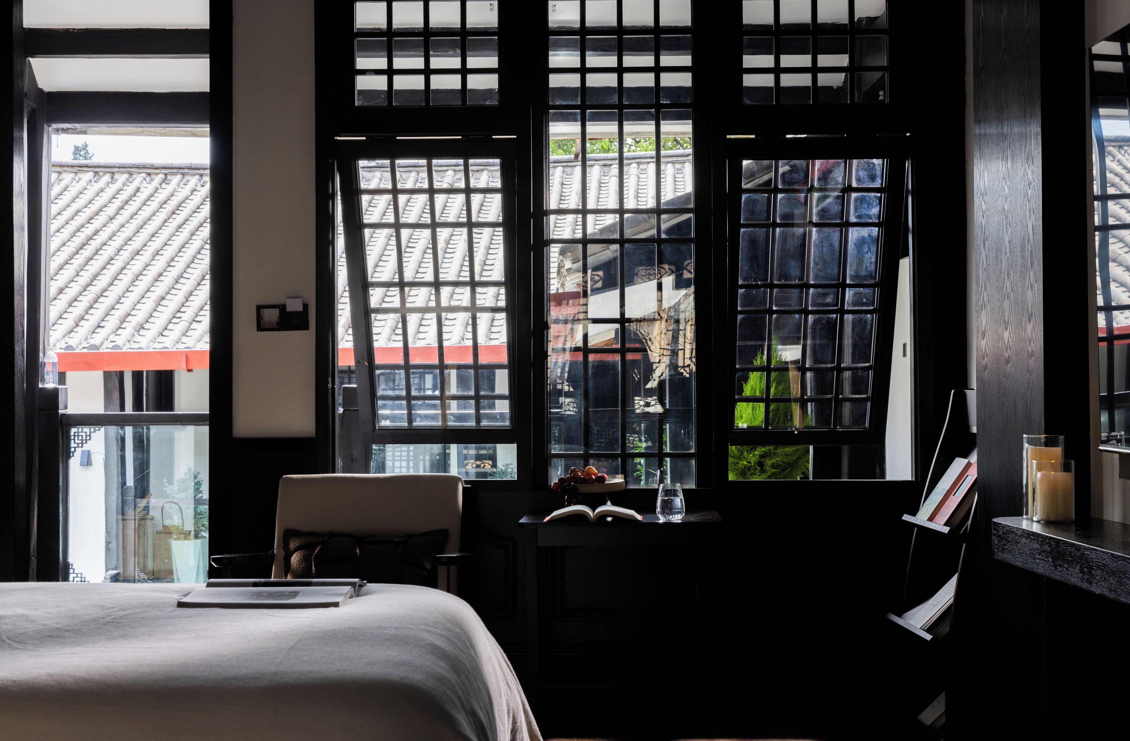 A hotel room with a bed, a desk with a chair, a window with a view of a traditional roof, and a small table with a candle and a glass.