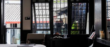 A hotel room with a bed, a desk with a chair, a window with a view of a traditional roof, and a small table with a candle and a glass.