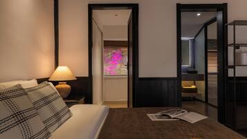 Une chambre d’hôtel avec un lit, une table de chevet avec une lampe, un magazine posé sur le lit et une vue sur une fleur rose à travers une porte ouverte.