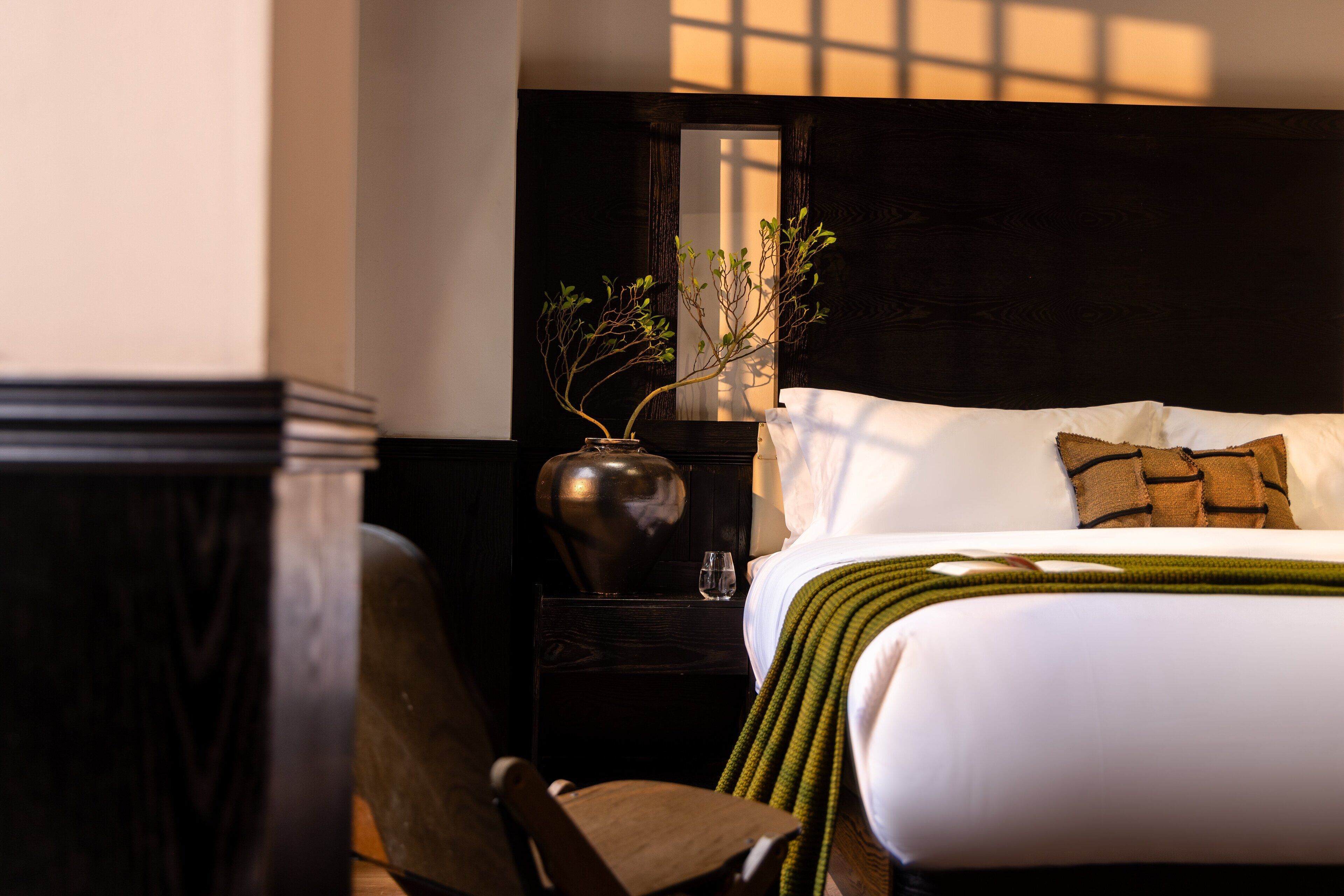Habitación de hotel impecable con una cama, una silla, una mesita de noche, un florero con planta y un espejo.