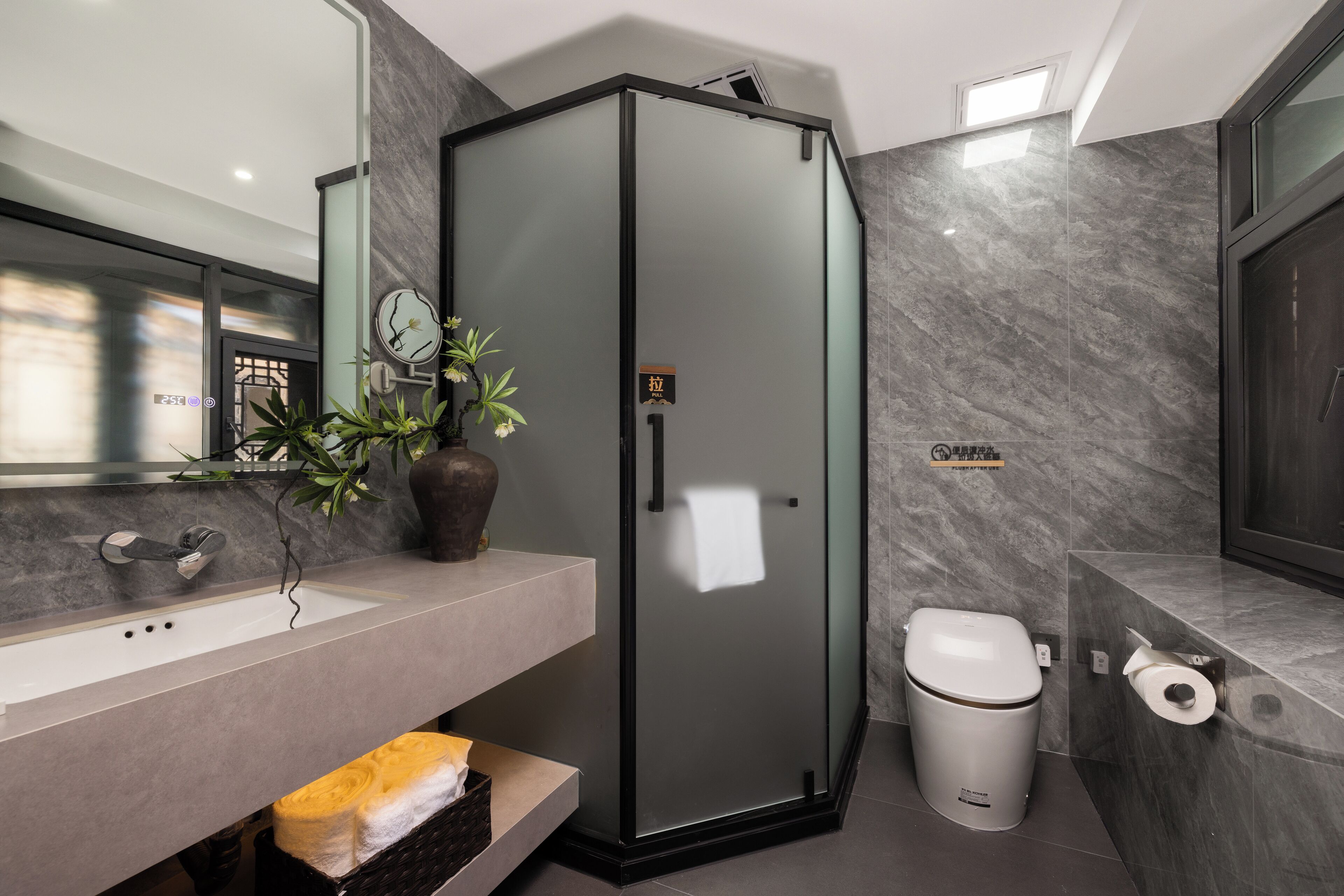 Een moderne badkamer met een glazen douchecabine, een wastafel met een bloemenvaas, en een toilet met een papieren handdoekdispenser.