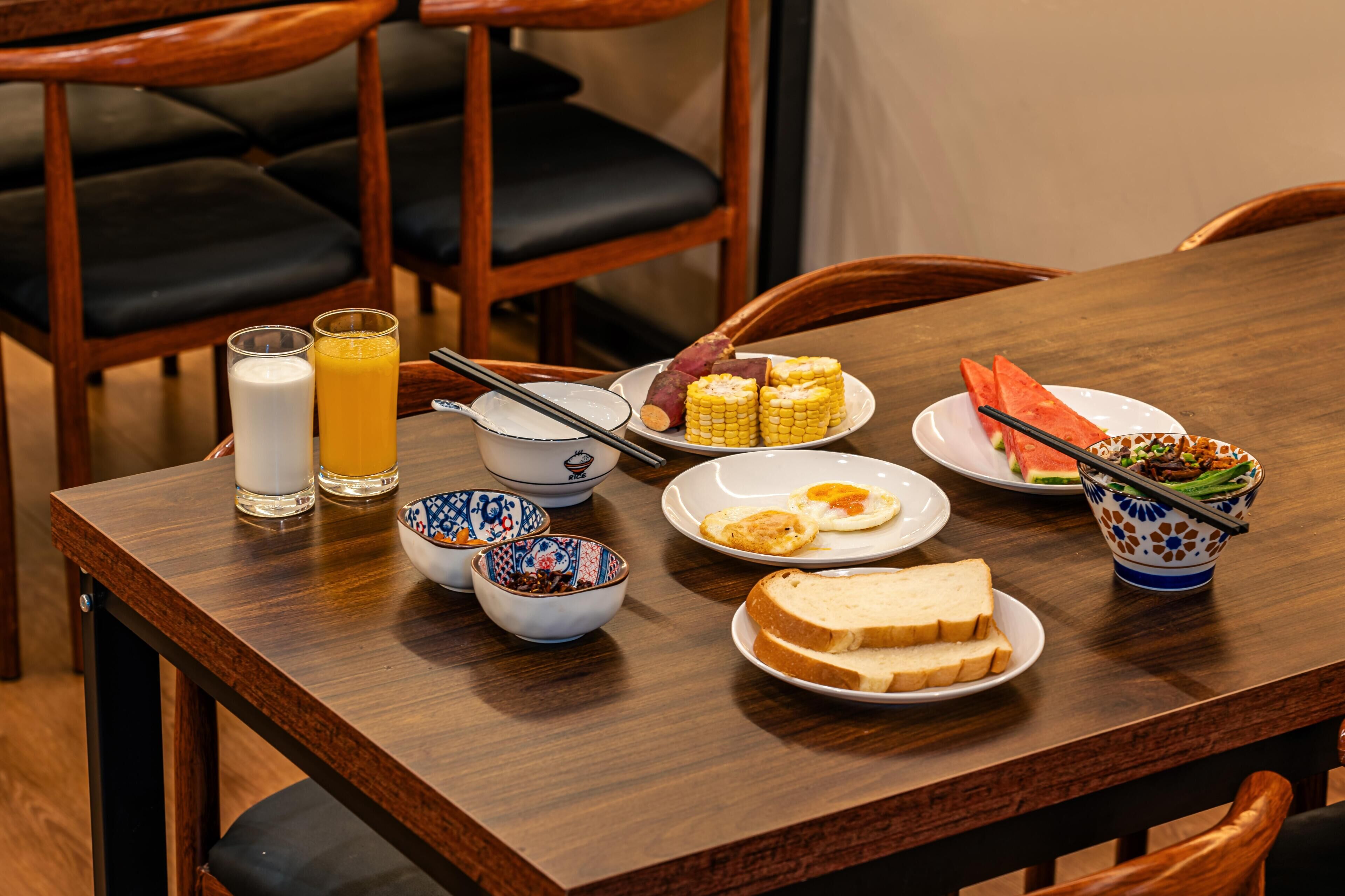 A wooden dining table with various dishes including corn on the cob, eggs, and watermelon, along with glasses of milk and orange juice.