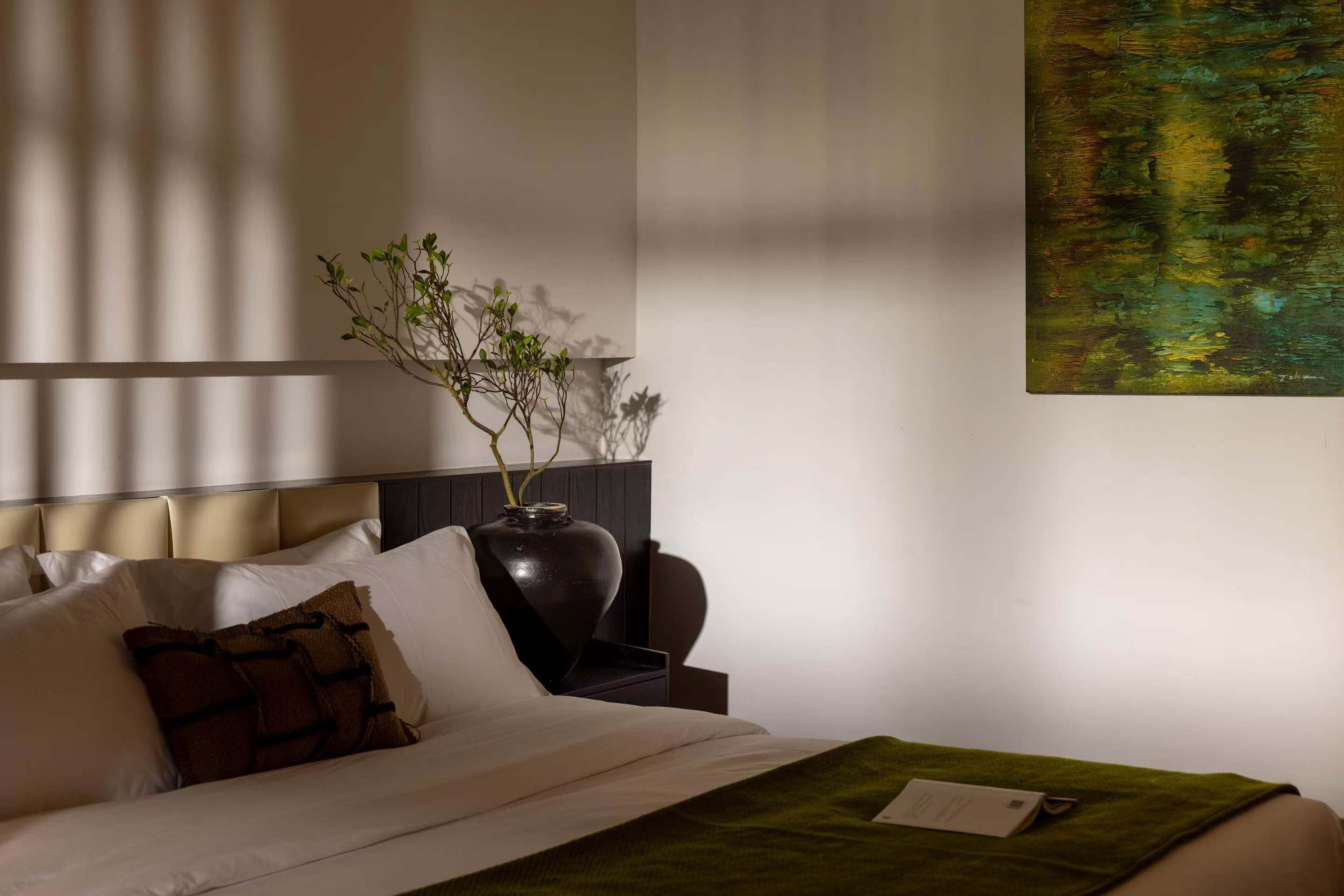 Ein Hotelzimmer mit Bett, Kissen, einer Vase mit Zweigen, einem Gemälde und einem Nachttisch.