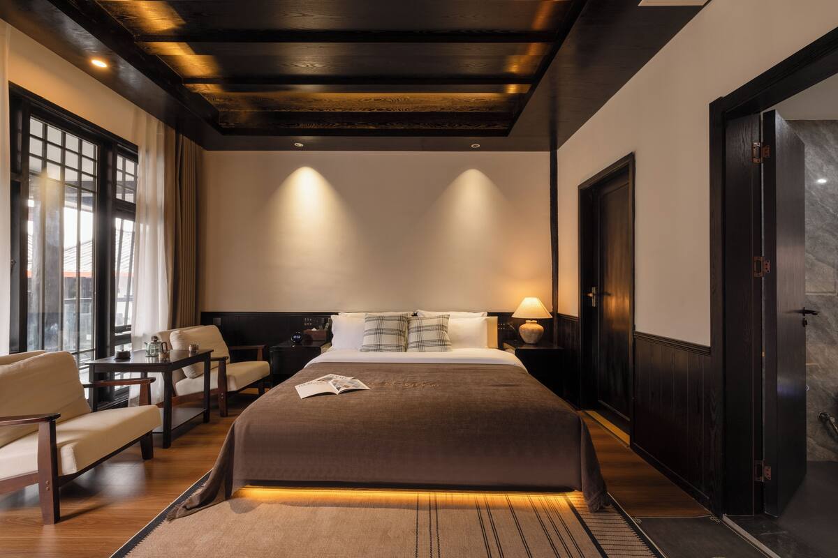 Una habitación de hotel moderna con una cama grande, un área de descanso con una mesa y sillas, y vista al exterior a través de una ventana.